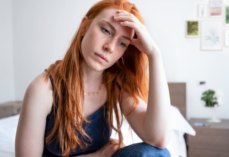 A concerned woman seated at the edge of her bed, with her hand resting on her forehead.