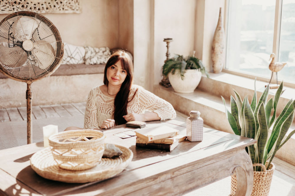 A woman seated on the floor of a bohemian-themed living room, with tarot cards and runes on the table.
