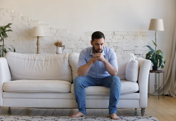 A young man seated on the couch in his living room, in deep thought.