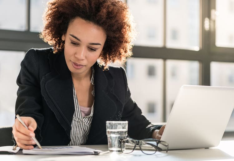 Woman sitting at a table with her laptop open, looking at a piece of paper.