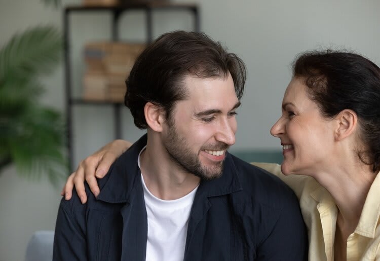 A young man wearing a white t-shirt, navy button-up shirt with a collar, with a dark brown slicked-back hair and some facial hair. Next to him, we see an older woman, wearing a pastel yellow button-up with a collar, with her hair tied back, faced towards the man, with her arm resting on his shoulder. Both of them have pleasant expressions. A bookcase and plant are faded out in the background.
