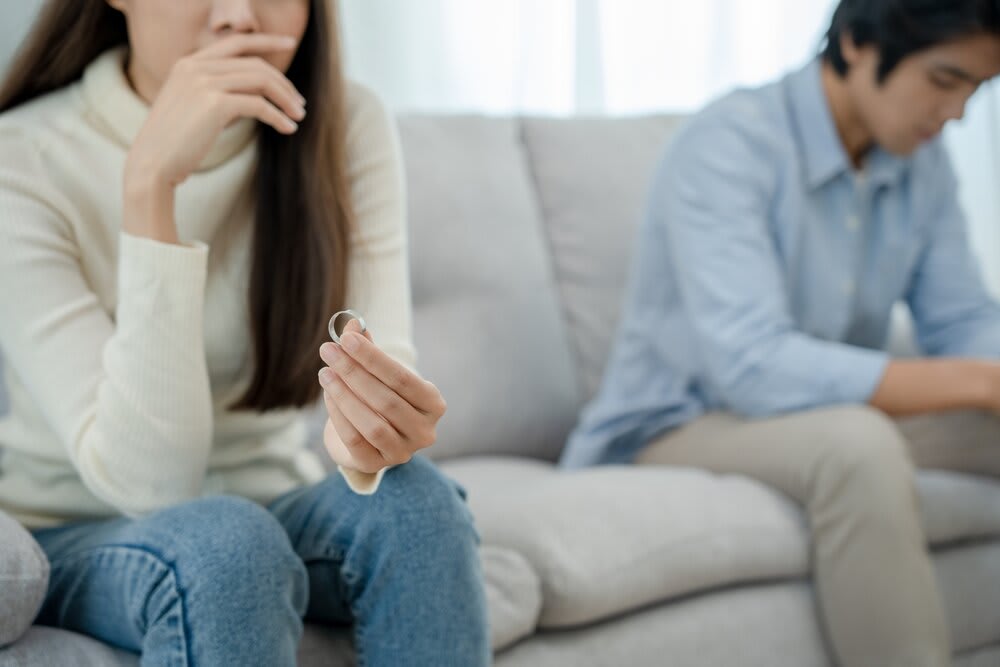A young couple seated on their couch, with the woman holding her wedding ring in her hand.