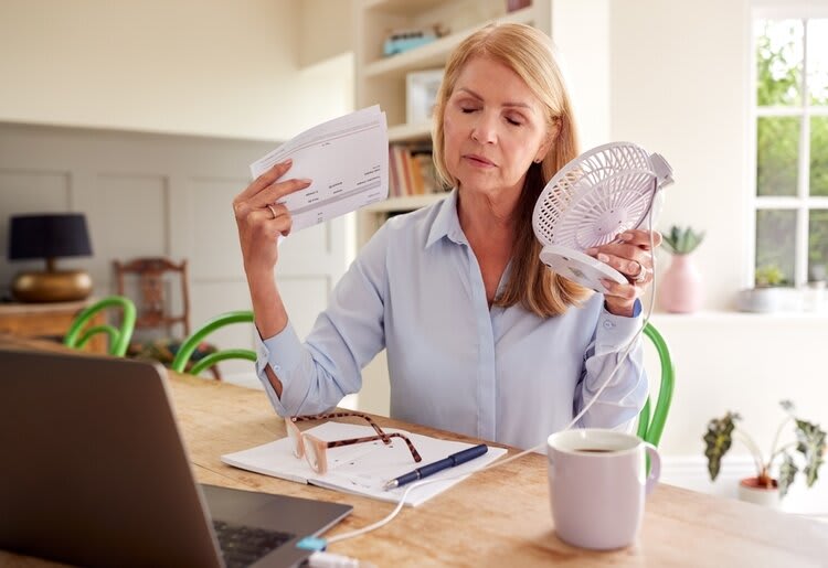 A mature woman experiencing a hot flush, cooling herself off with an electric fan and stack of papers.